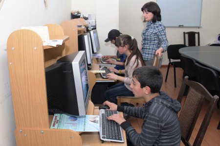 Drei Jugendliche sitzen vor den Computern, eine Lehrerin steht dabei.