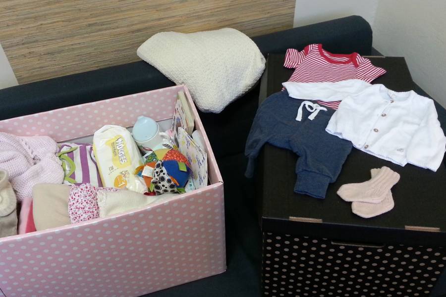 Ein Karton gefüllt mit einer Erstausstattung für ein Neugeborenes: Kleidung, Schuhe, Windeln, Trinkflasche.
