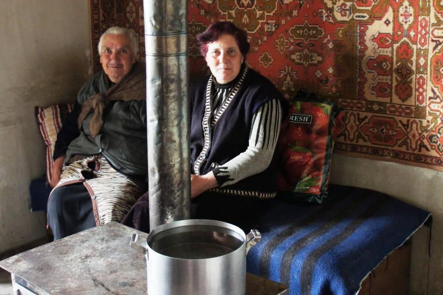 Zwei Frauen sitzen in einem Zimmer, davor steht ein Ofen, auf dem Wasser gekocht wird.