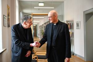 Kardinal Kurt Koch und Prof. Thomas Schwartz, Hauptgeschäftsführer von Renovabis, stehen in einem hellen Flur und geben sich die Hand. Beide Männer tragen schwarze Anzüge mit Priesterkragen. Im Hintergrund stehen Regale mit Büchern und es sind einige Stühle zu sehen. Die Wände sind weiß, an einer hängt ein Kalender und ein Bild. Der Flur ist gut beleuchtet und wirkt modern und freundlich.