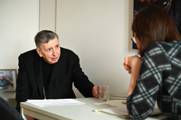 Kardinal Kurt Koch sitzt an einem weißen Tisch. Er trägt ein schwarzes Priestergewand. Vor ihm liegen Papiere. Er blickt aufmerksam auf Projektreferentin Christiana Hägele, die ihm gegenüber sitzt.