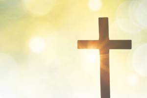 Auf einem weichen, hellgelben Hintergrund mit Lichtreflexen und angedeuteten Kreisen steht rechts im Bild ist ein schlichtes, dunkel silhouettiertes Kreuz, hinter dem warmes Licht hervorstrahlt – als Symbol für Hoffnung und Auferstehung.