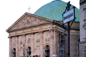 Fassade der Hedwigskathedrale in Berlin, danben steht ein Straßenschild mit der Aufschrift "Bebelplatz". Die Kathedrale hat fünf Bögen an der Frontseite, zwei davon sind zugemauert. Im Hintergrund eine grüne Kuppel.