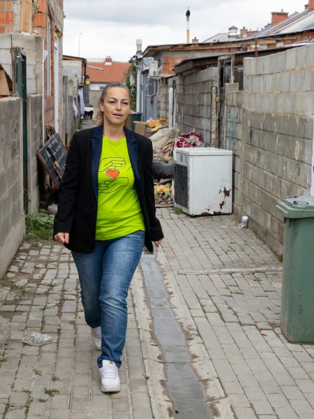 Edona Lipoveci, die Leiterin des Concordia-Tageszentrums in Gjakova, läuft durch eine Straße der Stadt. Sie trägt ein neongrünes T-Shirt, einen dunklen Blazer und mittelblonden Haare sind zum Zopf zusammengebunden. Auf der Straße liegt viel Müll und Unrat, die Häuser sind unverputzt.