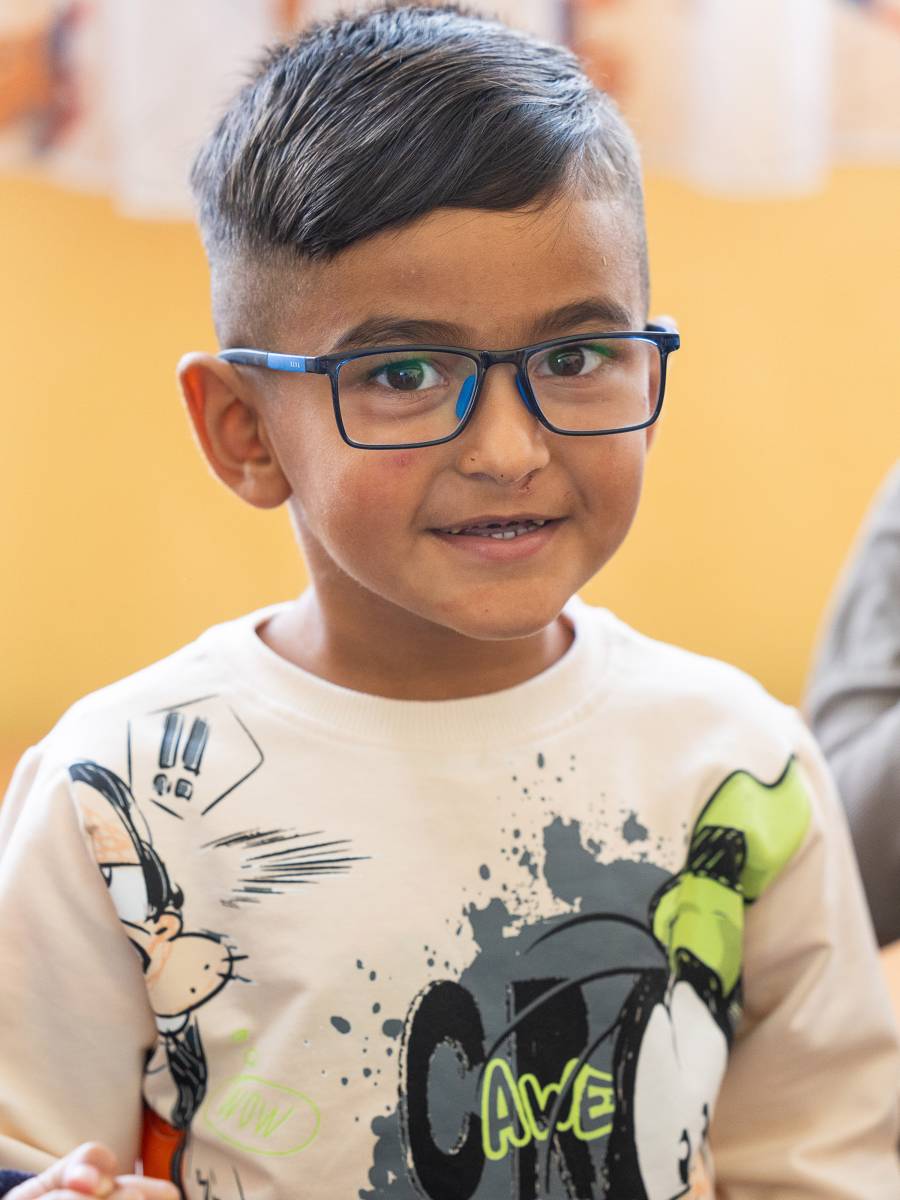 Ein kleiner Junge mit dunklen, glänzenden Haaren und einer großen dunklen Brille lächelt mit großen dunklen Augen etwas zaghaft in die Kamera des Fotografen.