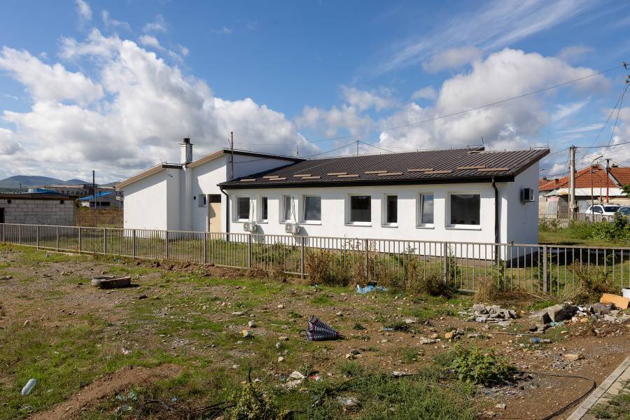 Das Gebäude des Ali-Ibra-Zentrums in Gjakova: ein schlichter weißer Zweckbau, eingeschossig, mit neuen Fenstern, das Gebäude und der Garten sind sehr gepflegt, doch außerhalb des Zaunes ist Müll und Unrat wie ein kaputter Regenschirm zu sehen.