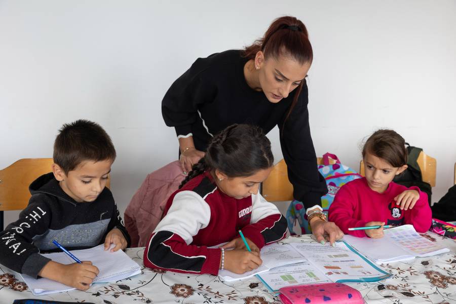 Eine Betreuerin und drei Kinder - zwei Mädchen und ein Junge - während des Nachhilfe-Unterrichts: Auf einer geblümten Tischdecke liegen aufgeschlagene Hefte, die Kinder halten Stifte in der Hand und schreiben gerade sehr konzentriert.