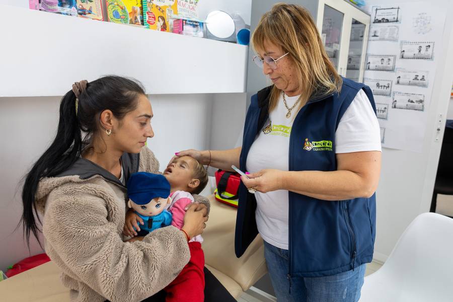 Eine junge Roma-Frau mit ihrem kranken, schlafenden Kleinkind wird in der Krankensprechstunde im Concordia-Zentrum in Malki Iskar von einer Mitarbeiterin betreut. Die Mitarbeiterin hält ein Fieberthermometer in der Hand.