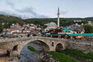 Das Bild zeigt die Altstadt von Prizren, im Vordergrund eine alte Stein-Brücke, dahinter Restaurants sowie eine Moschee mit einem Minarett, im Hintergrund sind bewaldete Hügel zu sehen.