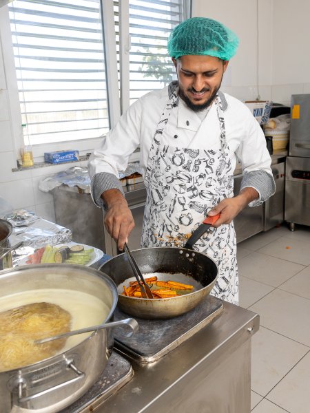 Der Koch Rahmi Mustafa steht am Herd einer Profi-Küche und rührt in einer Pfanne. Er trägt ein weißes Koch-Hemd und ein grünes Haarnetz.