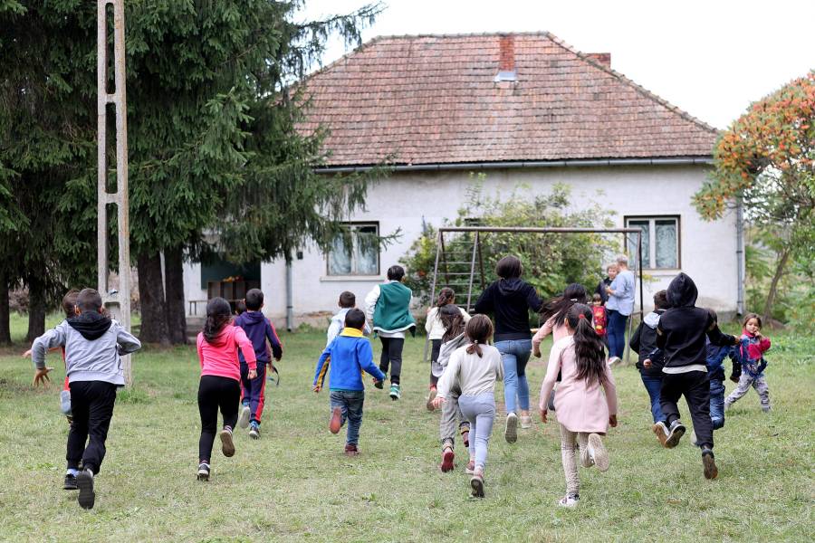 Mehr als ein Dutzend Kinder rennen vor ihrer Schule im Dorf Peteni über den grünes Gras auf ihr Schulgebäude zu. Im Hintergrund ist auch eine Schaukel zu sehen.