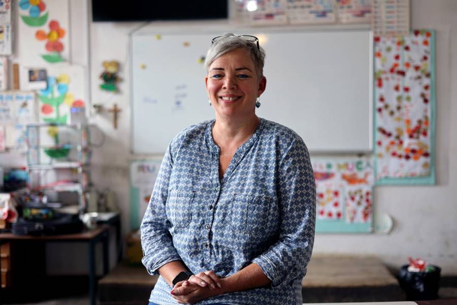 Porträtfoto der Lehrerin Anikó Haan Deàk im Klassenzimmer im Dorf Peteni in Rumänien. Sie hat kurze blonde Haare, ihre Brille hat sie auf den Kopf geschoben. Sie trägt eine blau-weiß gemusterte Bluse, im Hintergrund ist die Tafel zu sehen sowie einige Bilder und Basteleien der Schülerinnen und Schüler.