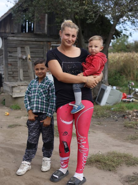 Eine junge Roma-Frau mit einem Kleinkind auf dem Arm und einem Schukind an ihrer Seite. Die Familie steht auf ungepflastertem Boden, im Hintergrund eine Holzhütte sowie ein kaputter Fernseher, der im Gras liegt.