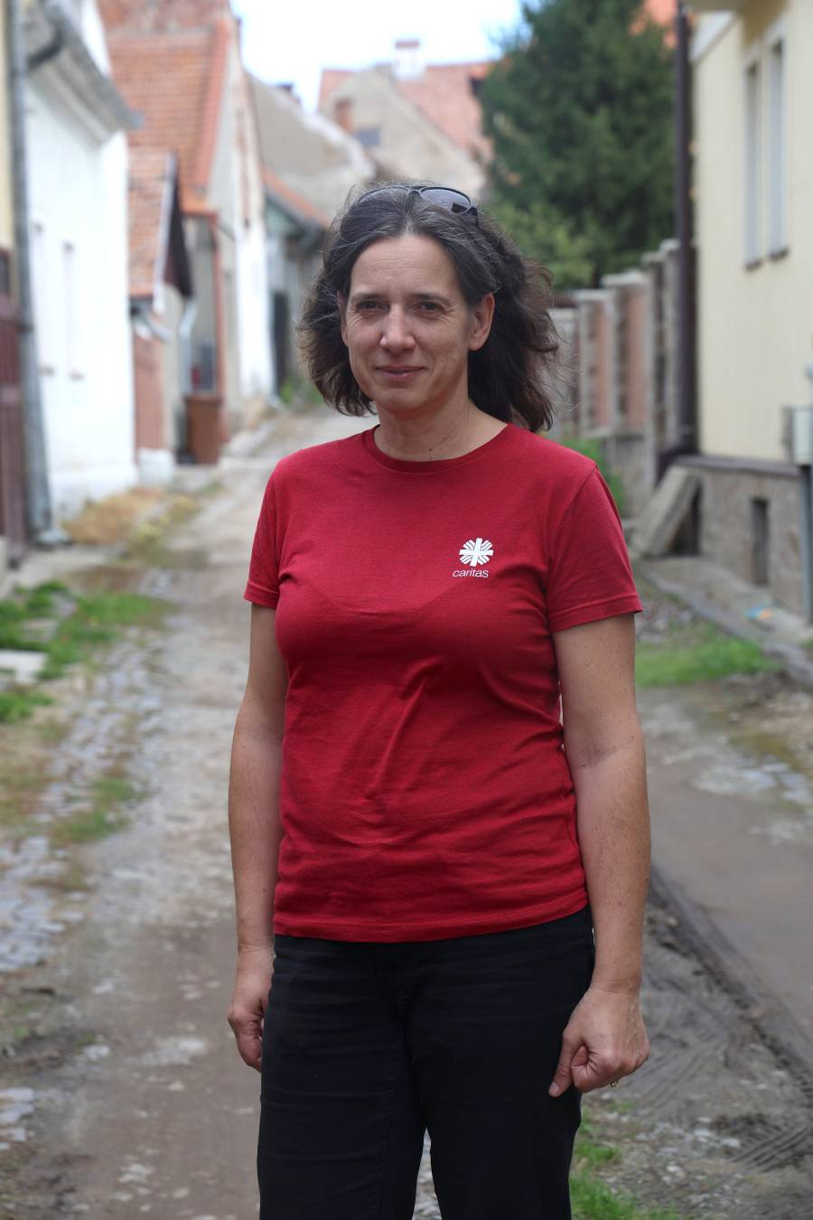 Kinga Hubbes, Koordinatorin der Caritas in Târgu Secuiesc, steht in einer schmalen Gasse. Sie trägt ein rotes T-Shirt mit der Aufschrift "Caritas" sowie eine schwarze Hose, ihre schulterlangen dunklen Haare trägt sie offen, die Brille hat sie in die Haare geschoben.