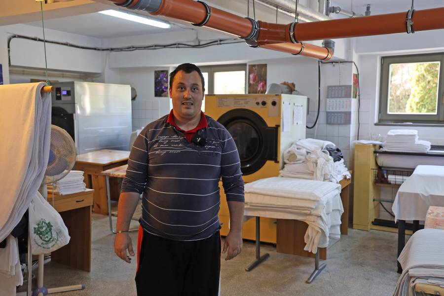 Ein Mann steht in einer Wäscherei vor Maschinen und schmutziger sowie sauberer Wäsche. Der Raum ist gefliest, an der Decke verlaufen graubraune Rohre.
