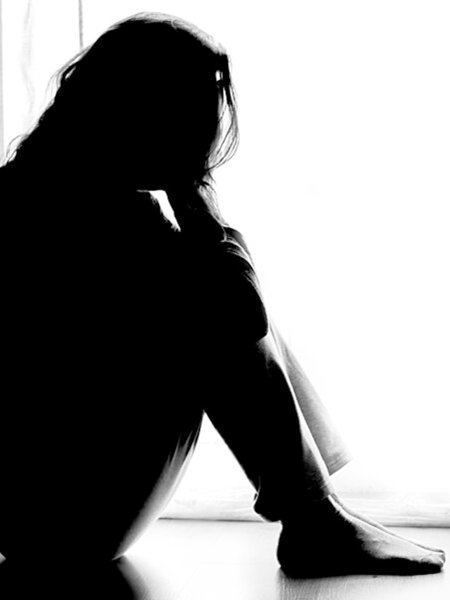 Schwarz-weiß-Aufnahme einer Frau im Gegenlicht. Sie sitzt mit angezogenen Beinen auf dem Boden und schlägt die Hände vor ihr Gesicht.