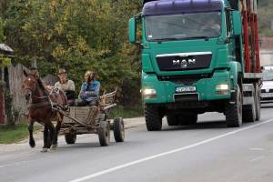 Auf einer geteerten Straße im ländlichen Rumänien fährt am rechten Fahrbahnrand ein Pferdefuhrwerk. Das Pferd zieht einen älteren Holzwagen, auf dem eine alte Frau und ein alter Mann sitzen. Dahinter naht ein großer, grüner Lastwagen.