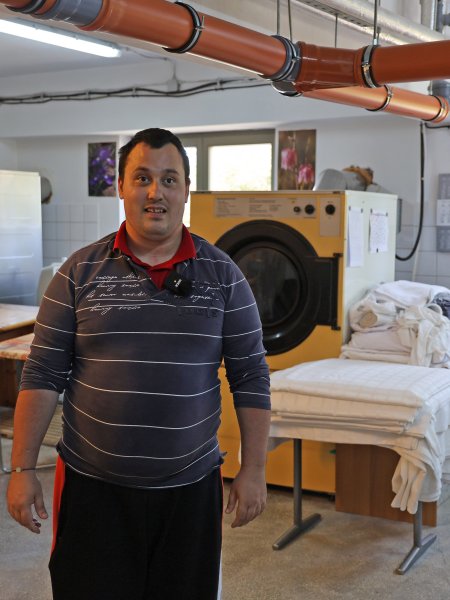 Loránt Péter steht in einer Wäscherei, um ihn herum riesige Waschmaschinen, Stapel von sauberer und auch schmutziger Wäsche. Er trägt ein graues Shirt mit weißen Streifen und eine dunkle Jogginghose.