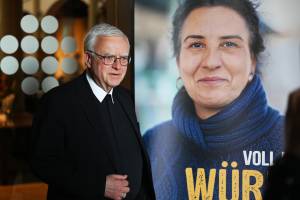 Erzbischof Heiner Koch steht neben einem Plakat zur Renovabis-Pfingstaktion 2025 mit dem Motto ‚Voll der Würde‘. Auf dem Plakat ist eine Frau mit dunklem Haar und blauem Rollkragenpullover abgebildet, die in die Kamera lächelt.