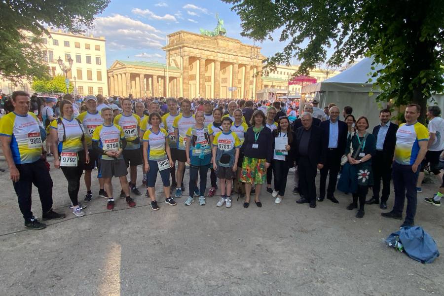 Eine Gruppe von Läuferinnen und Läufern von Misereor, Renovabis und dem Erzbistum Berlin, die sich vor dem Berliner Firmenlauf zu einem Gruppenfoto aufgestellt haben. Sie tragen weiß-gelb-blaue Lauftrikots mit Startnummer darauf.