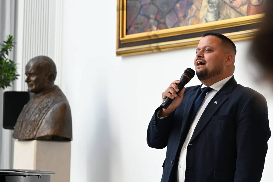 Das Bild zeigt Lukáš Bužo mit einem Mikrofon singend in der Berliner Nuntiatur. Er ist Abgeordneter im slowakischen Parlament und MItglied im slowakischen Musik-Ensemble F6. Er trägt einen dunklen Anzug, ein weißes Hemd und eine dunkelblaue Krawatte. Er hat kurze, dunkle Haare und trägt einen dunklen Kinn- und Schnauz-Bart.