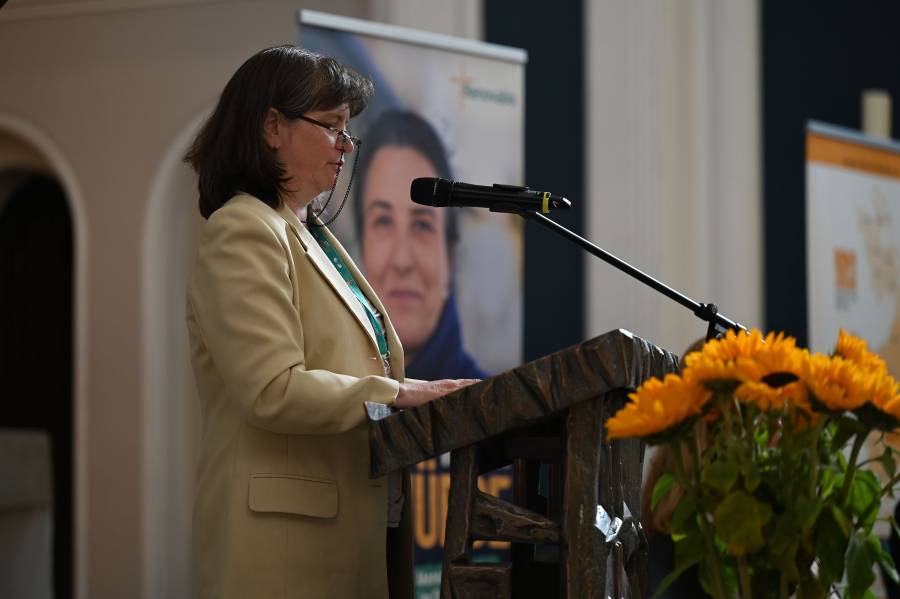 Erika Klemm, Migrationsbeauftragte aus Rumänien, steht in einem beigen Blazer, mit dunklen, halblangen Haaren und einer Brille mit dunkler Brillenkette am Mikrofon in der Zwölf-Apostel-Kirche in Berlin. Vor ihr steht ein Strauß leuchtend gelber Blumen.