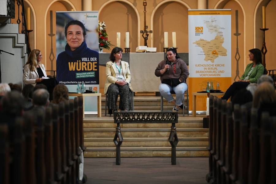 Das Bild zeigt die Diskussionsteilnehmerinnen der Podiumsveranstaltung „Die Würde der Frau ist (un)antastbar!?" in der Berliner Zwölf-Apostel-Kirche. Die vier Frauen sitzen auf Metallstühlen im Altarraum der Kirche, eine spricht gerade in ein Mikrofon. Rechts und links hängen Plakate von Renovabis und dem Erzbistum Berlin.
