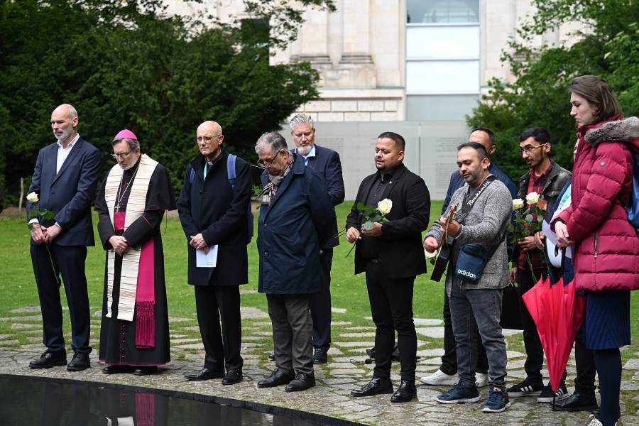Eine Gruppe von dunkel gekleideten Frauen und Männern steht am Denkmal für die im Nationalsozialismus ermordeten Sinti und Roma Europas in Berlin. Sie halten weiße Rosen in den Händen.