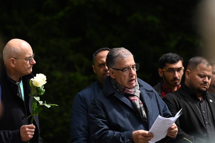 Mehrere Männer stehen in Jacken nebeneinander. Der Mann links hält eine weiße Rose in der Hand. Der Mann in der Mitte blickt auf ein paar weiße Papiere in seinen Händen, während er eine Rede hält.