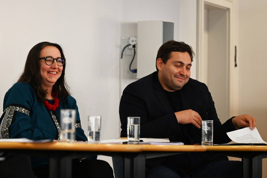 Das Bild zeigt eine Frau, links, und einen Mann, rechts, an einem Tisch sitzend. Beide lächeln.