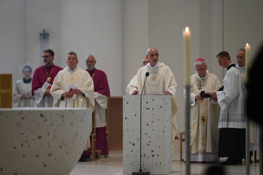 Das Bild zeigt eine Szene in der Sankt Hedwigs-Kathedrale in Berlin. Zu sehen ist ein weiß gekleideter Mann an einem Rednerpult. Hinter ihm sind weitere Männer in priesterlichen Gewändern zu sehen.