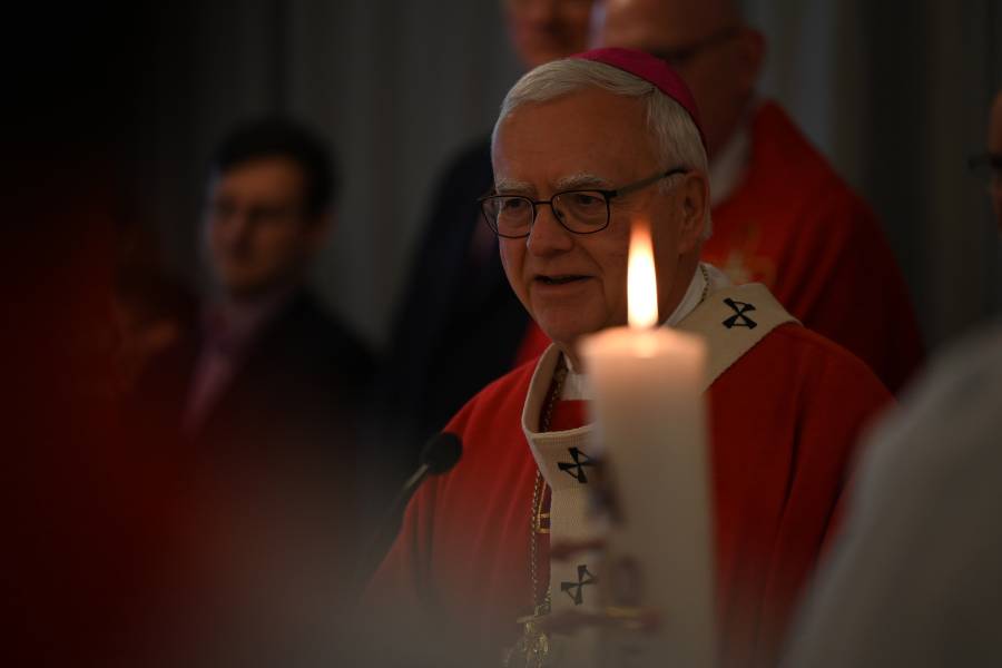 Im Vordergrund des Bildes ist leicht unscharf eine brennende Kerze zu sehen, im Hintergrund der Berliner Erzbischof Dr. Heiner Koch im roten Messgewand inmitten von Gläubigen.