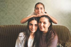 Drei Frauen – Mutter und Töchter – sitzen in inniger Pose auf einem gemusterten Sofa vor einer schlichten Wand. Während die jüngere Tochter mit einem schelmischen Lächeln zur Seite blickt, schauen die beiden anderen Frauen lächelnd direkt zum Betrachter. Die Szene vermittelt ein starkes Gefühl von Geborgenheit und familiärer Solidarität.