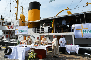 Ein Gottesdienst unter freiem Himmel. Im Hintergrund liegt ein Dampfschiff an, das mit Renovabis-Plakaten behängt wurde.