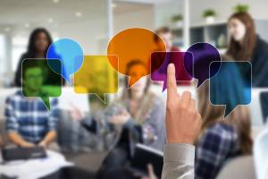 The photo depicts a group of people engaged in a discussion or meeting. The scene is overlaid with colorful, semi-transparent speech bubbles in various shapes, symbolizing communication, exchange of ideas, or dialogue.