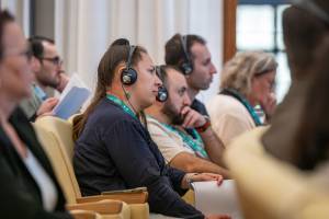 Mehrere Kongressteilnehmende sitzen im Publikum und tragen Kopfhörer für die Simultanübersetzung. Sie verfolgen aufmerksam die Vorträge und halten Tagungsunterlagen in den Händen.