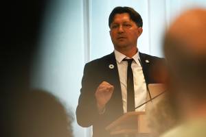 Ein Mann mit Anzug steht an einem Rednerpult, im Vordergrund sind die Zuhörenden zu erkennen.