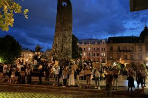 Das Bild zeigt einen Platz in der Ukrainischen Stadt Lviv. Es ist bereits dunkel. Auf dem Platz stehen viele Menschen, die Schilder in die Höhe halten, auf denen Sätze auf ukrainisch geschrieben sind.