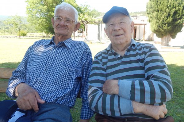 Zwei alte Männer sitzen im Garten auf einer Bank und schauen in die Kamera.