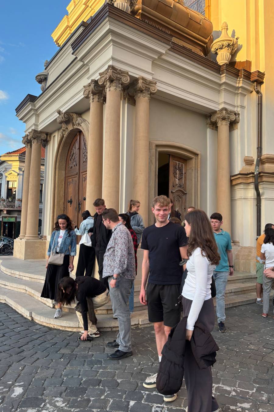 Junge Frauen und Männer stehen vor dem Gebäude einer Kirche. Das Gebäude ist gelb, davor der Eingangsbereicht mit Säulen und drei Treppenstufen.