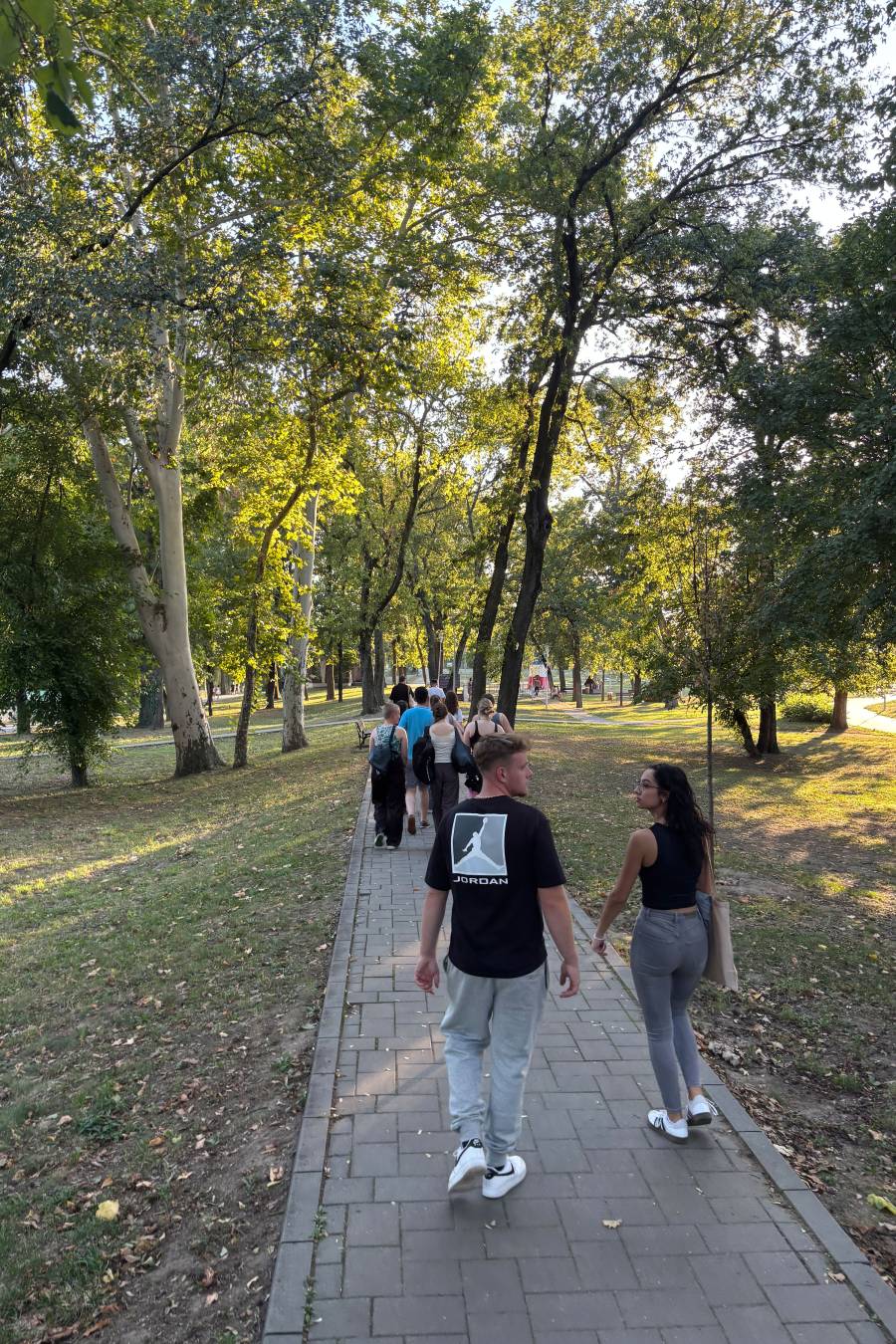 Jugendliche auf dem Weg durch einen Park. Sie tragen Jeans und laufen auf einem schmalen geteerten Weg unter saftig-grünen Bäumen hindurch.