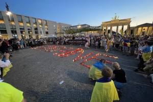 Das Bild zeigt den Pariser Platz vor dem Brandenburger Tor. Darauf sind viele Menschen zu sehen, die sich ukrainische Fahnen umgehängt haben. In der Mitte des Platzes ist das Kerzen das Wort "Ukraine" gebildet.