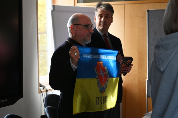Militärbischof Bohdan Manyschyn hält die Fahne des Militärseelsorge-Departements der Ukrainischen Griechisch-Katholischen Kirche hoch. Sie ist wie die ukrainische Fahne blau-gelb. Darauf ist ein rotes Emblem abgebildet.