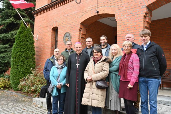 Das Bild zeigt die Reisegruppe, bestehend aus 10 Personen und den Erzbischof von Riga, Dr. Zbignevs Stakevičs vor einem Backsteinhaus, an dem die Flagge Lettlands weht.