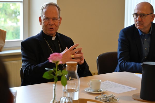 Das Bild zeigt zwei Männer, die an einem Tisch sitzen. Links ist der Erzbischof Dr. Gintaras Grušas zu sehen, der gerade spricht und die Hände vor sich faltet.