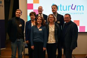 Das Bild zeigt eine Gruppe Menschen, die gemeinsam auf einer Bühne stehen. Im Hintergrund ist auf einem Bildschirm das Wort "Futurum" zu lesen.
