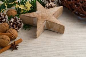 Adventliche Dekoration auf hellem Untergrund: Im Zentrum liegt ein fünfzackiger Stern aus naturbelassenem Holz. Drumherum sind Walnüsse, Zimtstangen, Sternanis und kleine Tannenzapfen arrangiert, teilweise mit weißem Reif überzogen. Grüne Tannenzweige und gelblich geränderte Stechpalmenblätter rahmen das Arrangement ein. Am rechten Bildrand ist ein geflochtener Kranz aus Weidenruten zu sehen.