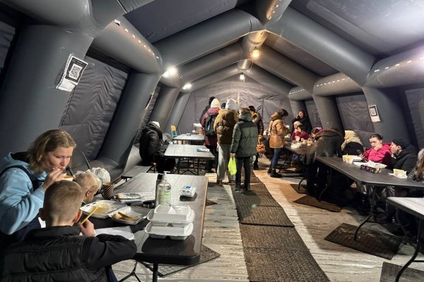 In einem großen grauen Zelt sitzen Menschen in Winterjacken an Tischen und essen.