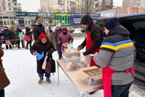 Das Bild zeigt eine Schlange Menschen, die in dicken Winterjacken im Schnee anstehen, um an einem Tisch Lebensmittel zu erhalten.