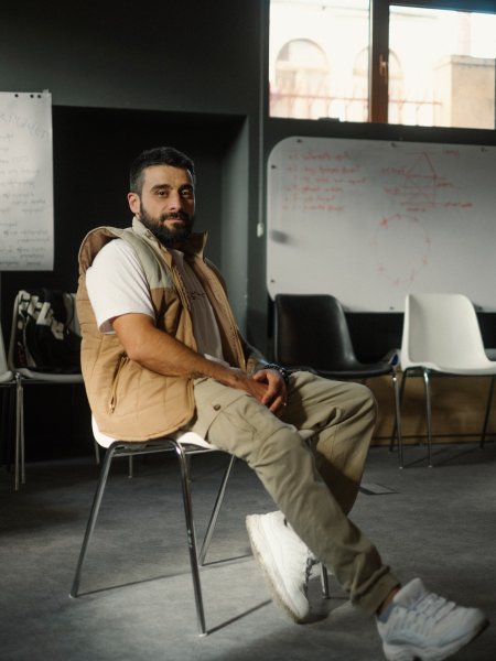 Der Rapper Lyoka aus Armenien. Er sitzt mit beiger Hosen, hellem Shirt und einer ärmellosen Jacke auf einem Stuhl.